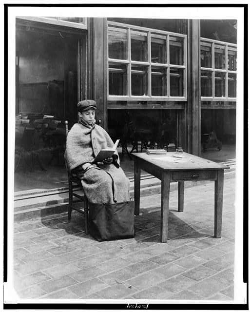 A student in a "sitting-out bag" sitting outdoors at a desk, reading, between 1900 and 1920.